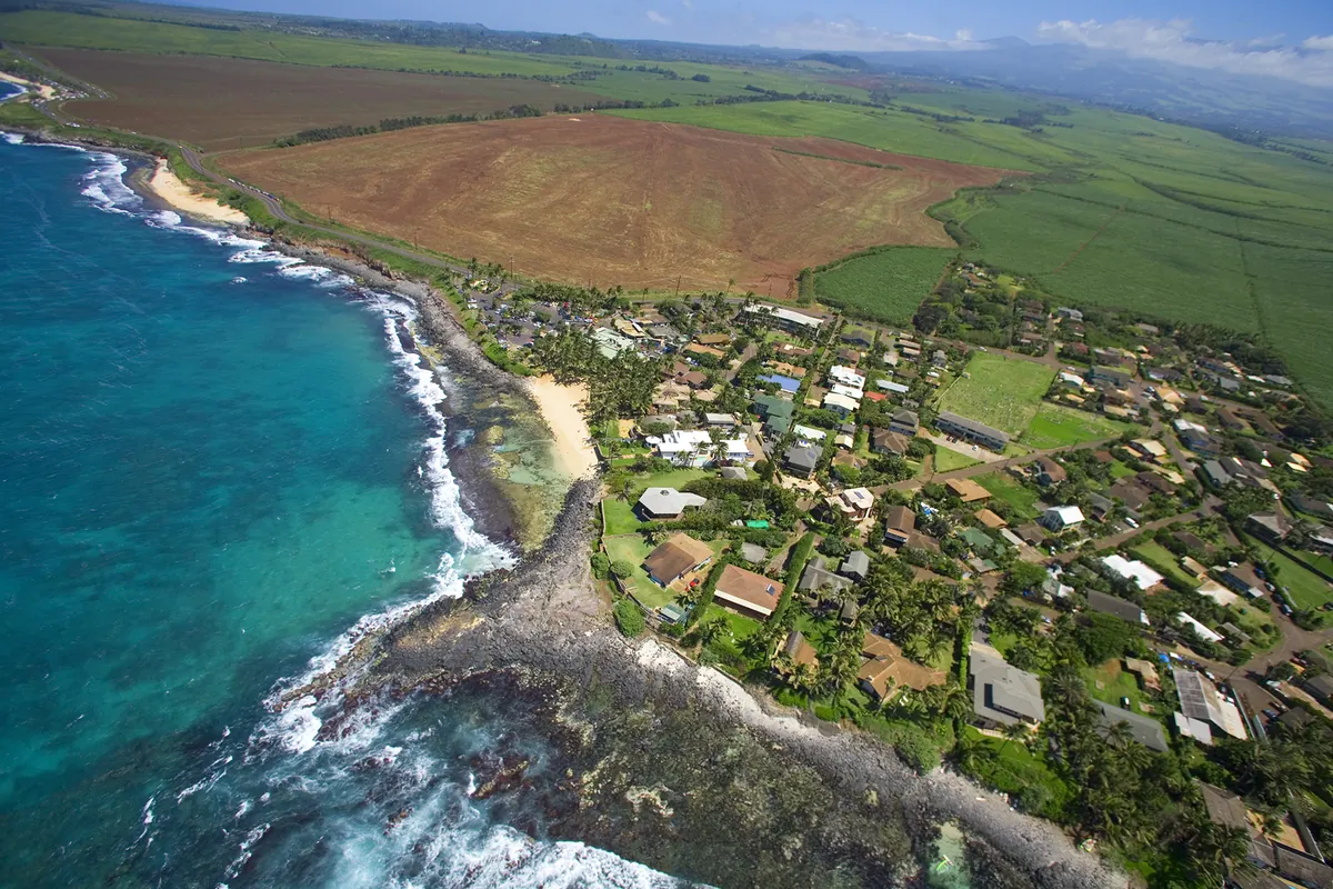 Aerial view of a Maui north shore neighborhood on a rocky point, turquoise water on one side and green fields rising inland.